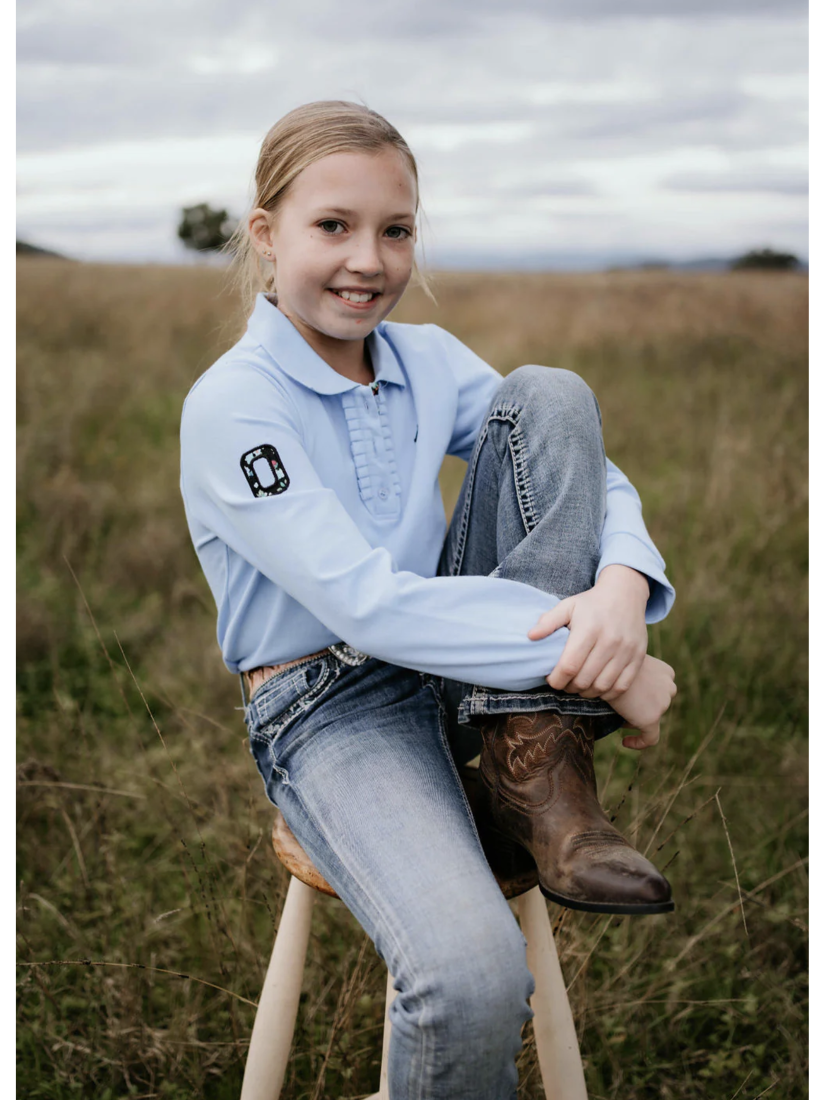 Young girl sitting on a stool in a field wearing a light blue shirt and jeans with cowboy boots.