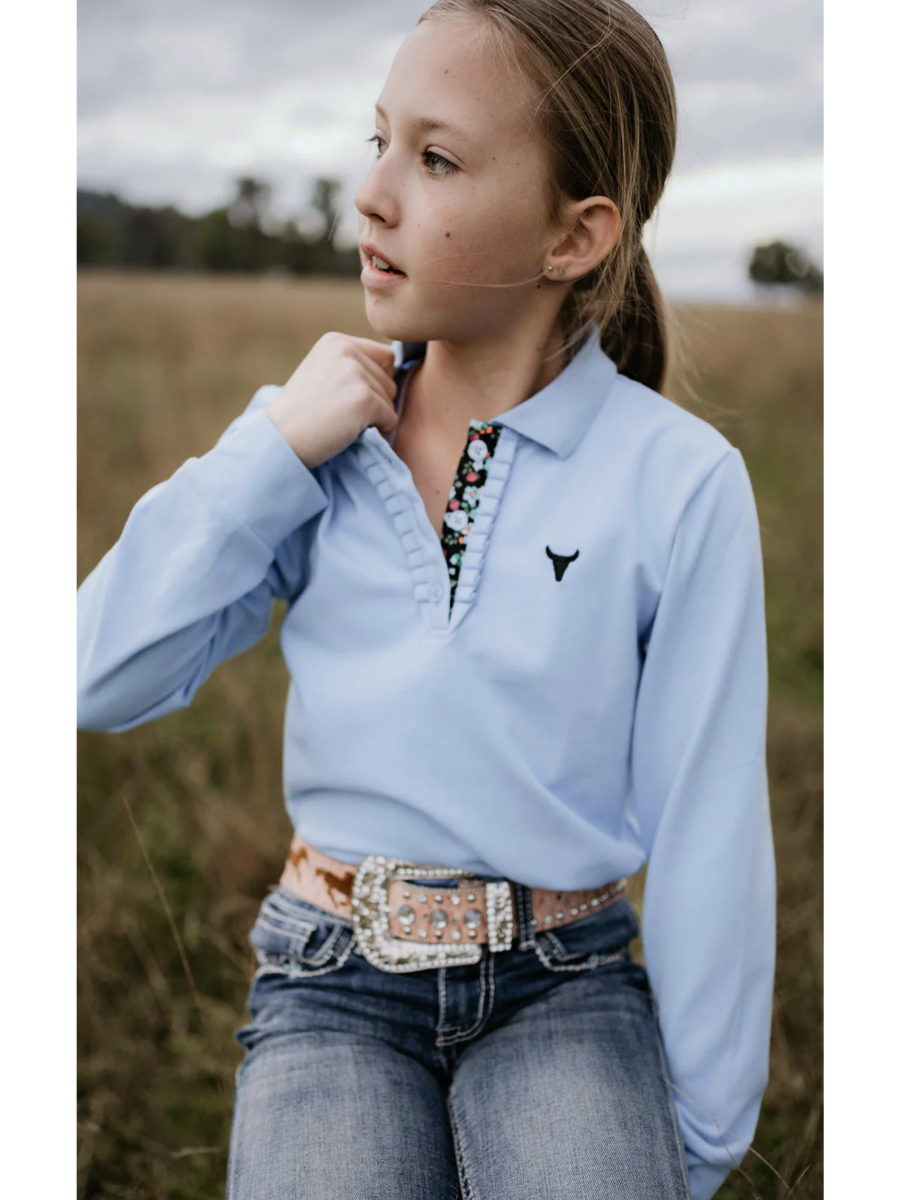 Girl wearing a light blue long-sleeve shirt with a logo, jeans, and a belt in a field.