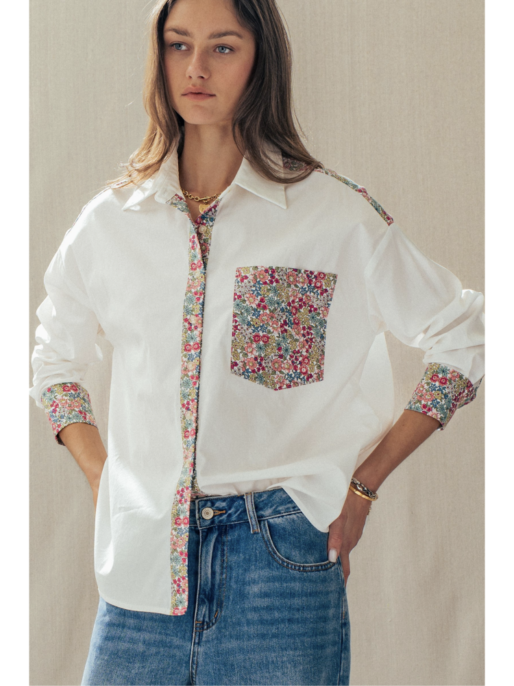 Woman wearing a white shirt with floral accents and blue jeans against a neutral background