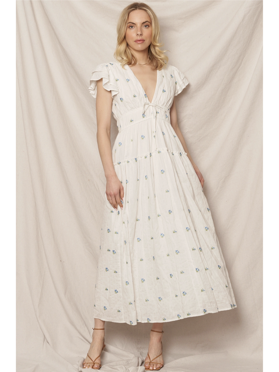Woman in a white tiered maxi dress with a blue and peach floral pattern, featuring flutter sleeves, standing against a clean white background.