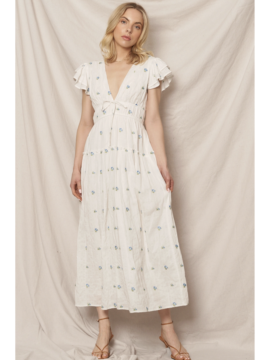 Woman in a white tiered maxi dress with a blue and peach floral pattern, featuring flutter sleeves, standing against a clean white background.