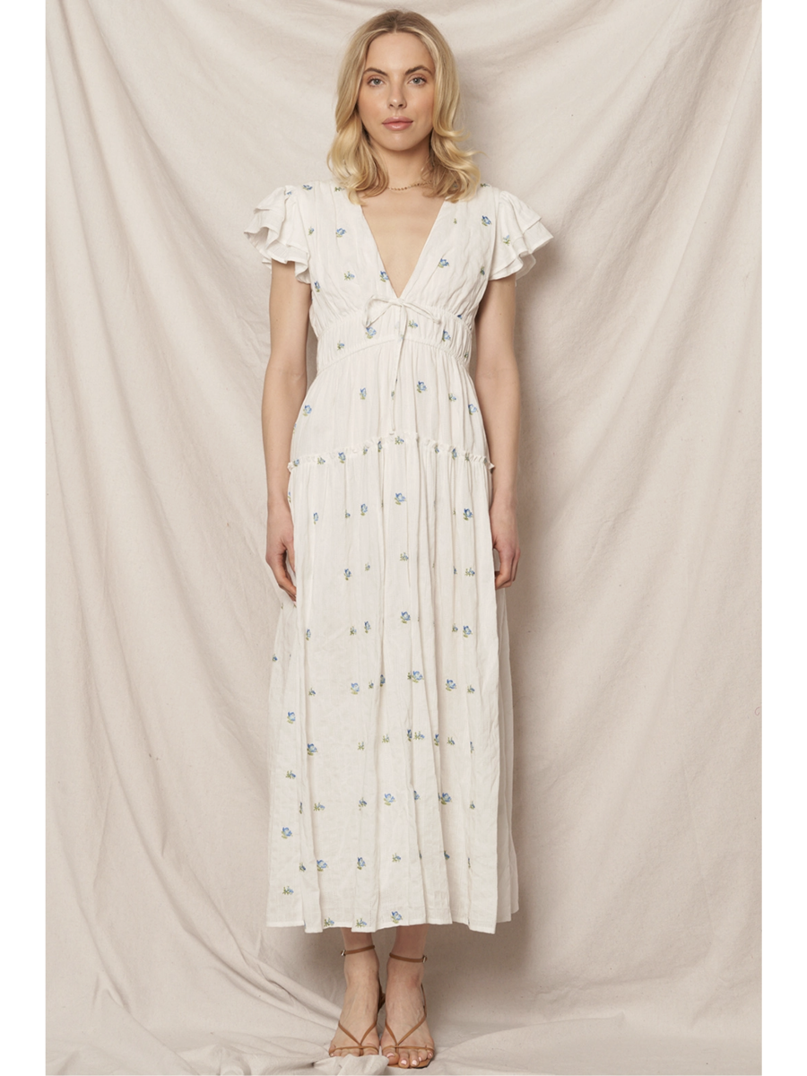 Woman in a white tiered maxi dress with a blue and peach floral pattern, featuring flutter sleeves, standing against a clean white background.