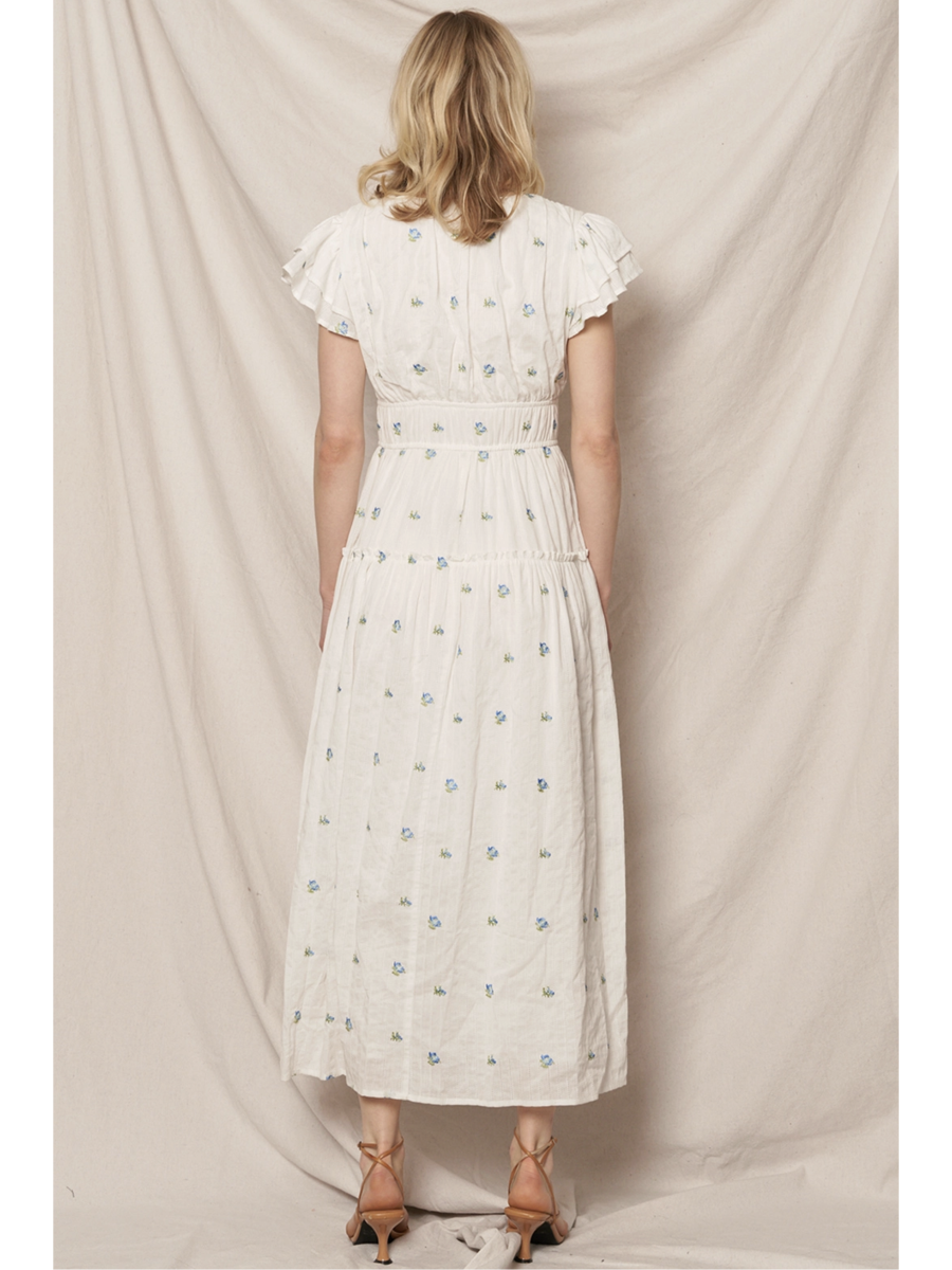 Woman in a white tiered maxi dress with a blue and peach floral pattern, featuring flutter sleeves, standing against a clean white background.