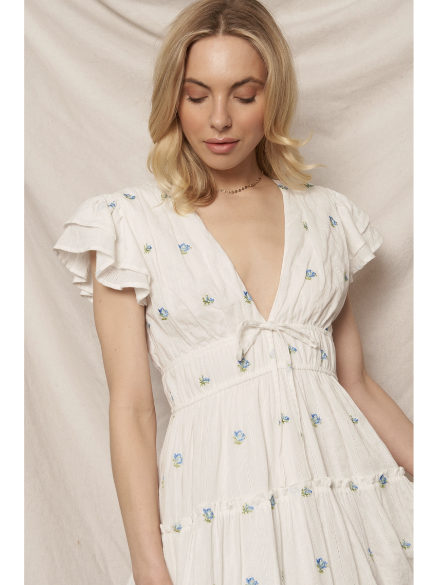 Woman in a white tiered maxi dress with a blue and peach floral pattern, featuring flutter sleeves, standing against a clean white background.