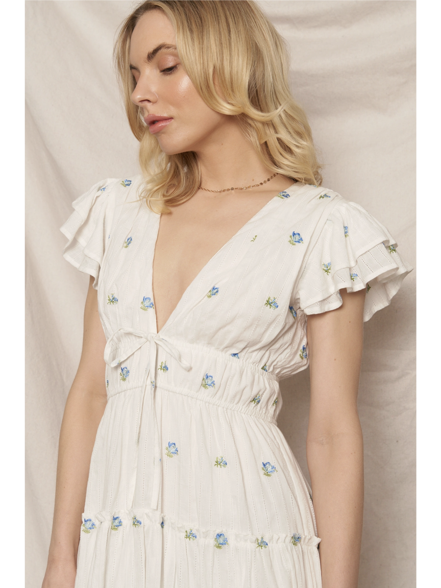 Woman in a white tiered maxi dress with a blue and peach floral pattern, featuring flutter sleeves, standing against a clean white background.