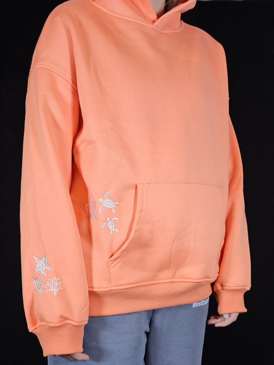 Person wearing a peach hoodie with white embroidered designs of sea life on a black background