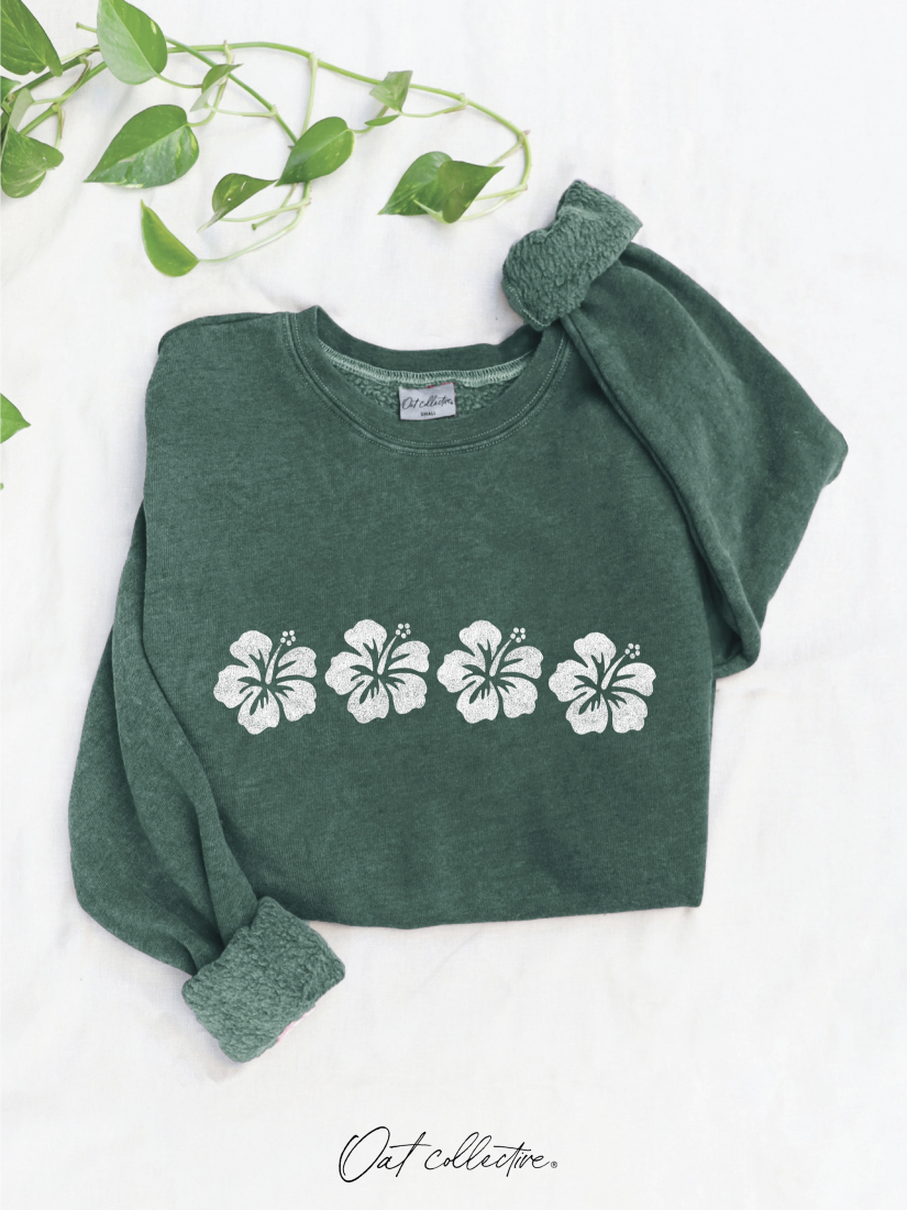 A dark green crewneck sweatshirt with four white hibiscus flowers printed across the chest, styled flat on a white fabric background with a green trailing plant in the top-left corner.