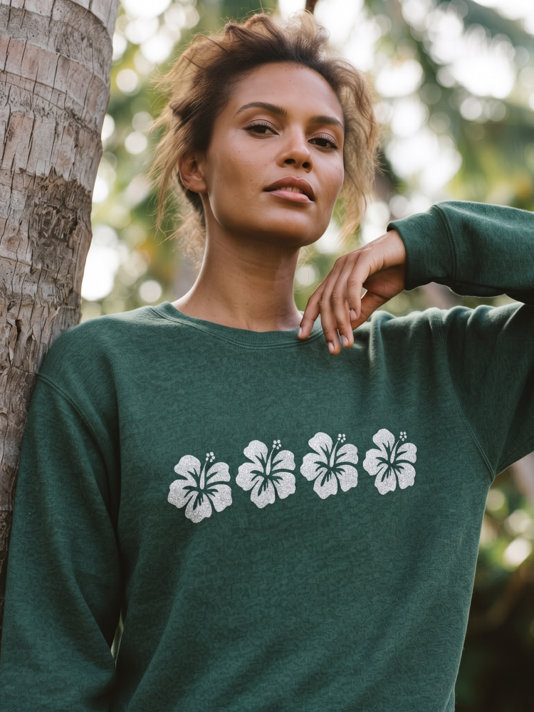 A woman wearing a dark green crewneck sweatshirt with a white hibiscus flower graphic across the chest, posing outdoors next to a tree trunk.