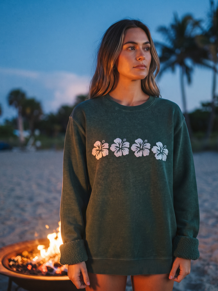 A woman stands on a beach at dusk wearing an oversized dark green sweatshirt with a horizontal row of five white hibiscus flowers across the chest. A fire pit with flames is visible in the foreground, and palm trees and sand are in the background.