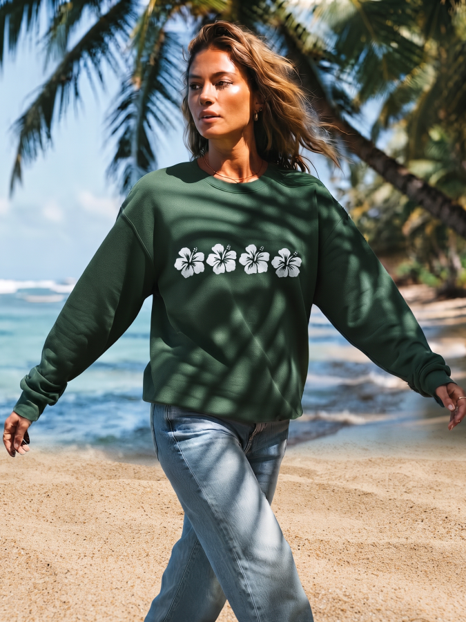 A woman walking on a sandy beach wearing a dark green crewneck sweatshirt with a white hibiscus flower graphic across the chest, paired with light blue jeans.