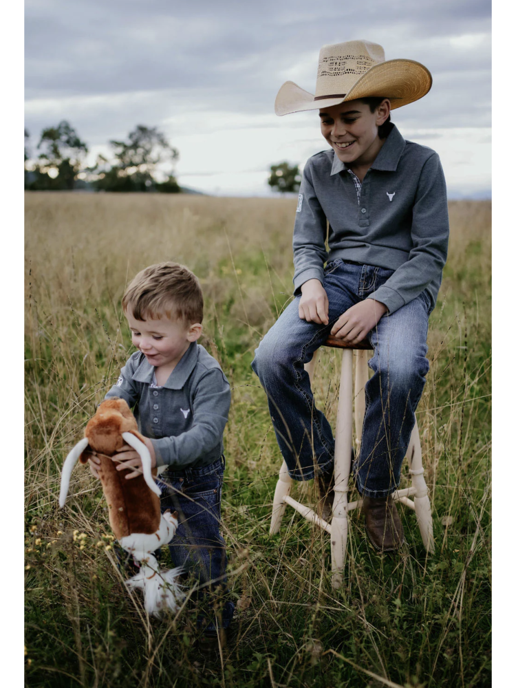 Two children in a field, one sitting on a stool wearing a cowboy hat and the other holding a stuffed animal.