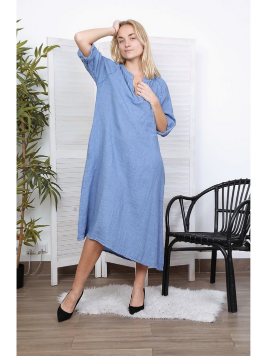 Woman wearing a sky blue 100% linen long dress with a V-neck and relaxed fit, standing on a white rug next to a black chair.