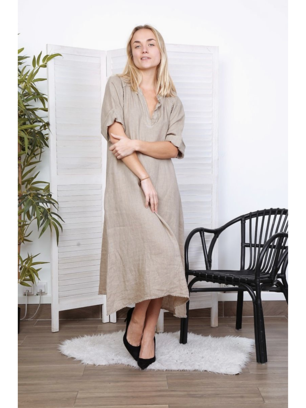 A woman wearing a long taupe 100% linen dress with ruffled sleeves and a V-neck, standing in a bright room with a white room divider.