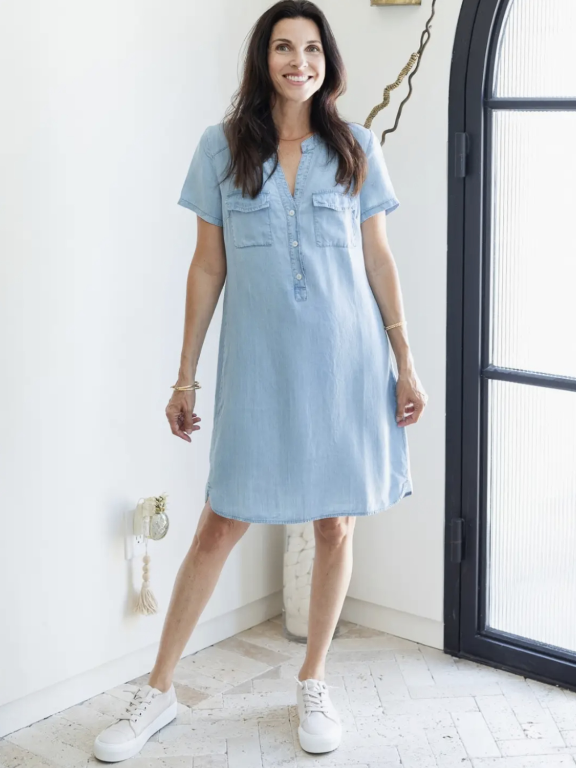 A woman modeling a light blue denim short-sleeve button-front dress with two chest pockets, paired with white sneakers, standing in a bright indoor room with a white wall and a black-framed window.