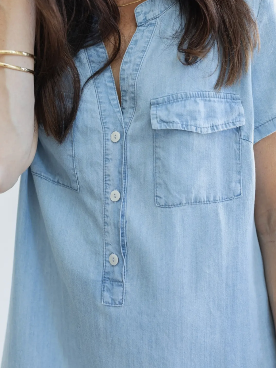 A close-up of a person wearing a light blue chambray short-sleeve shirt dress with a button-down front and a chest pocket.