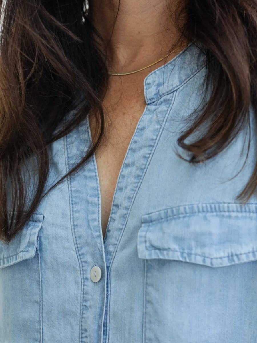 Close-up of a person wearing a light blue denim button-up shirt with chest pockets and a small gold chain necklace.