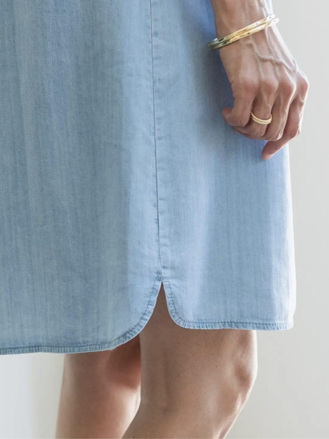 Close-up of a person wearing a light blue chambray dress, showing the side slit and hand with gold bangles and a ring against a plain light background.