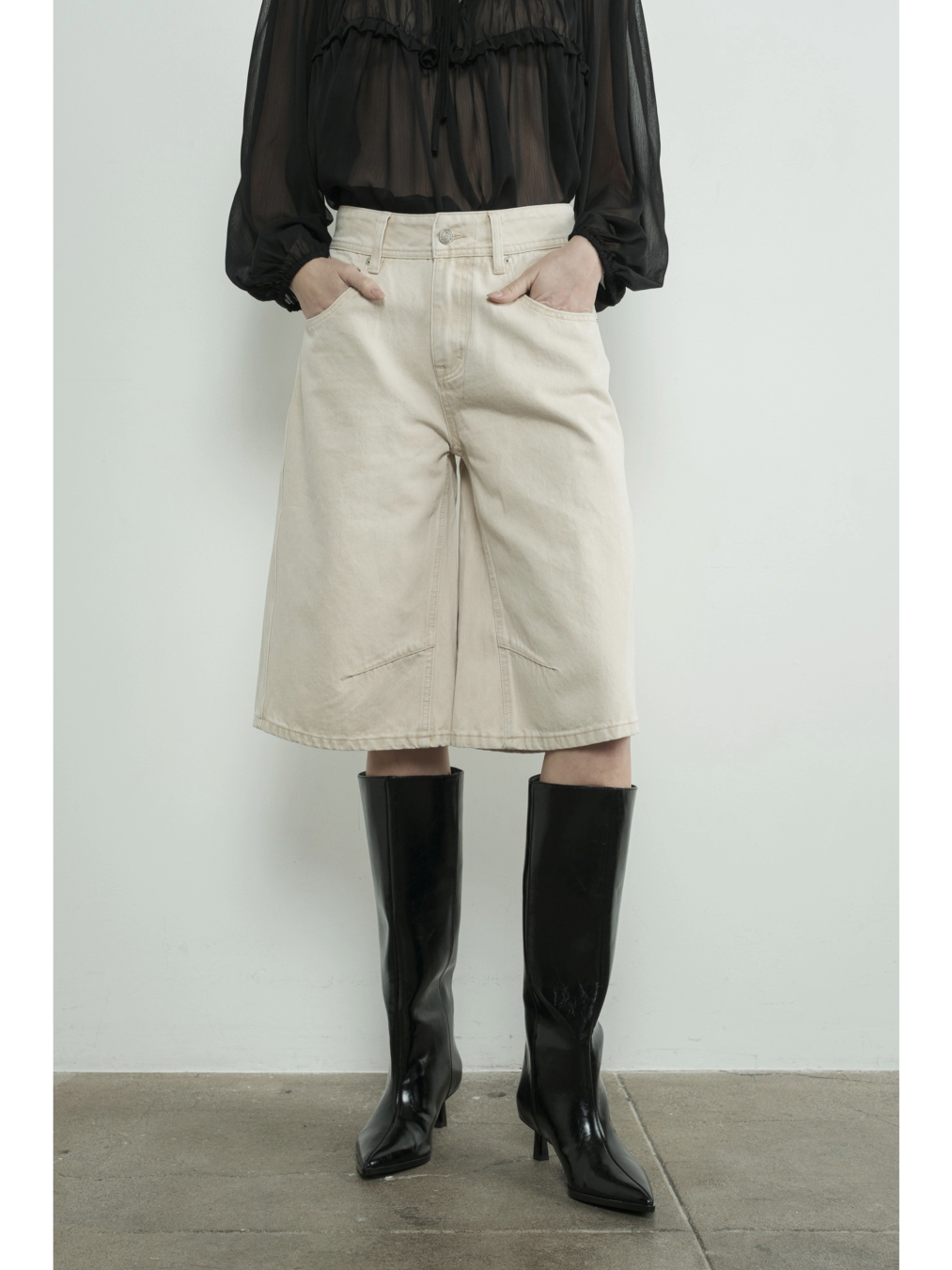 A woman wearing high-waisted cream Bermuda shorts with a yoke seam detail and side pockets, paired with a white top against a neutral background.