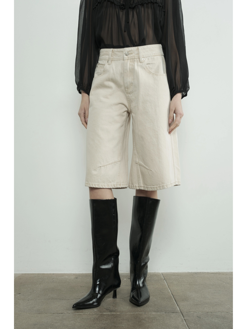 A woman wearing high-waisted cream Bermuda shorts with a yoke seam detail and side pockets, paired with a white top against a neutral background.
