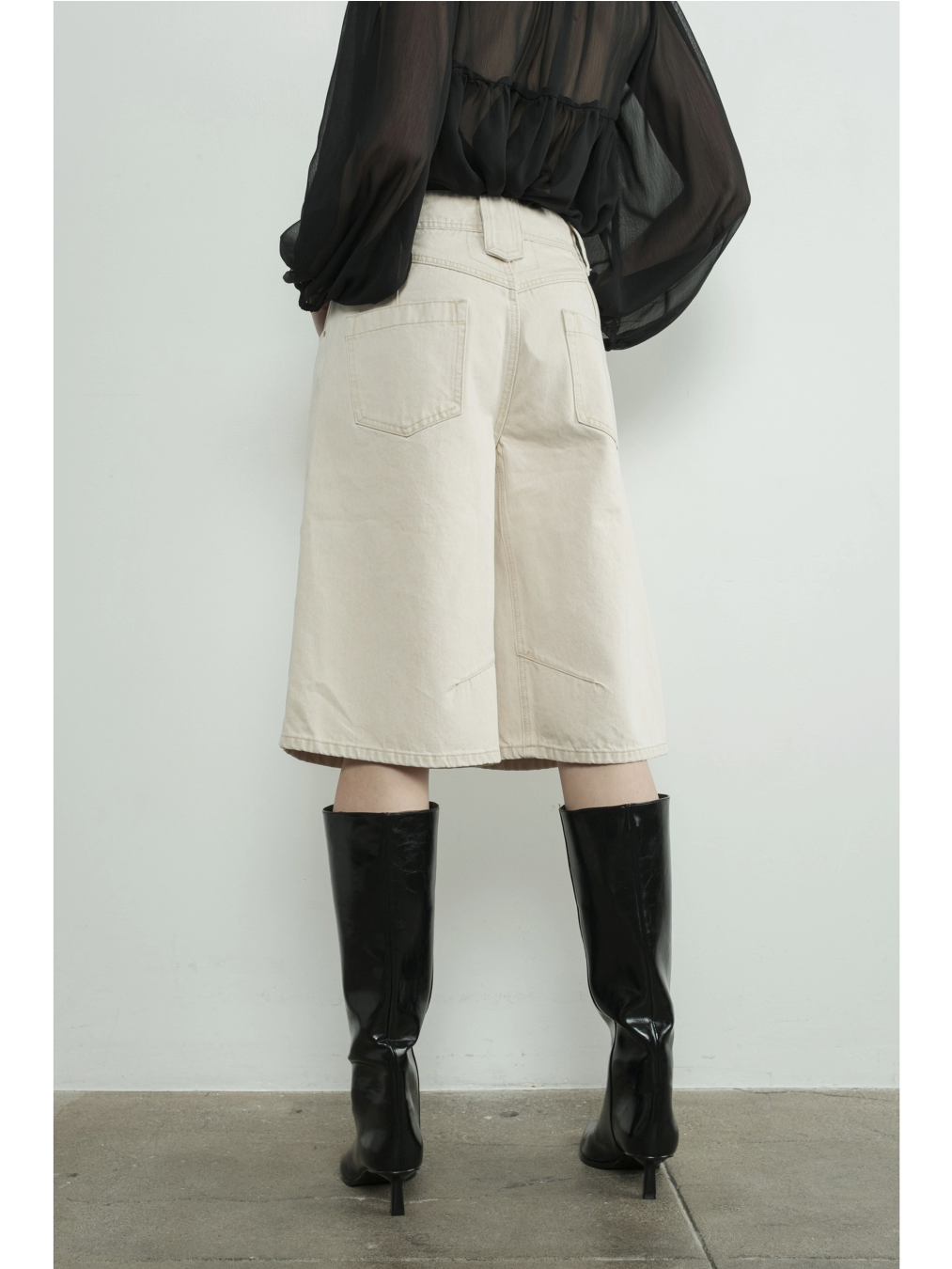 A woman wearing high-waisted cream Bermuda shorts with a yoke seam detail and side pockets, paired with a white top against a neutral background.