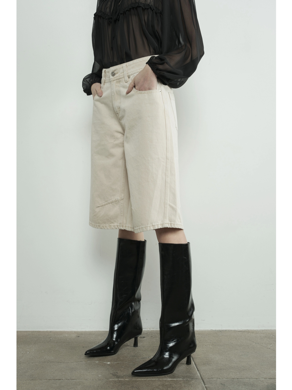A woman wearing high-waisted cream Bermuda shorts with a yoke seam detail and side pockets, paired with a white top against a neutral background.