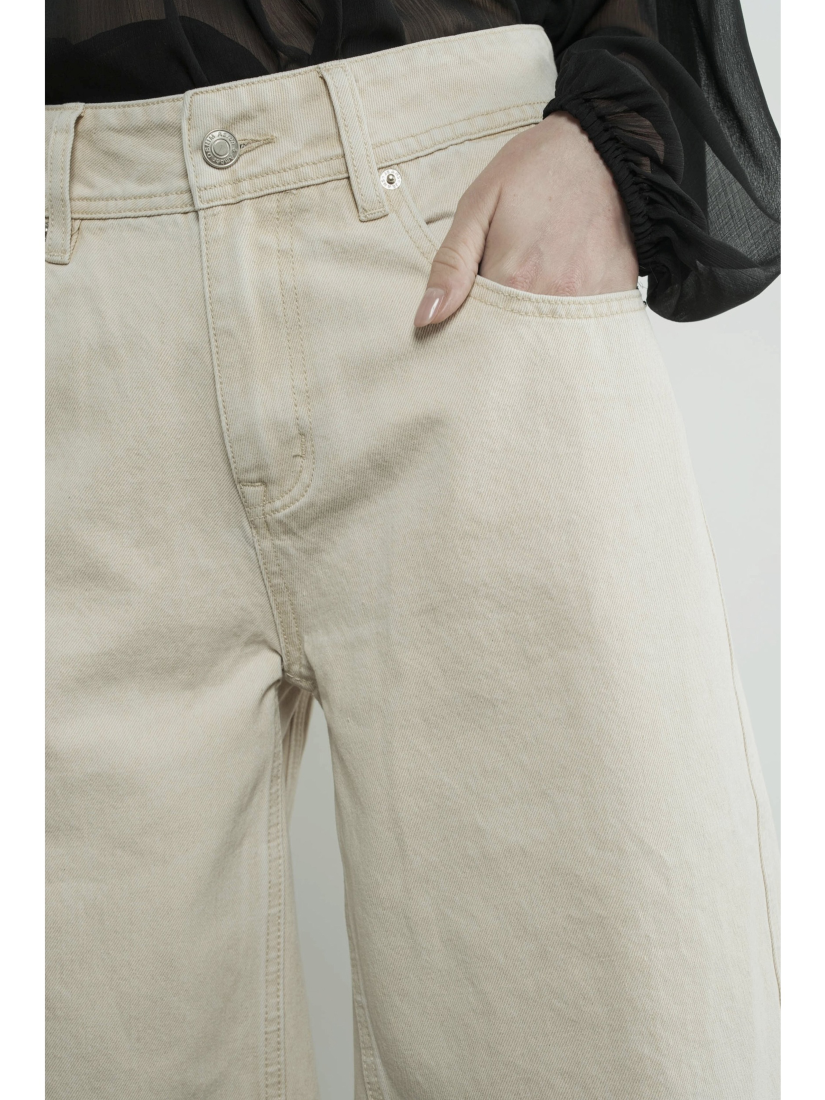 A woman wearing high-waisted cream Bermuda shorts with a yoke seam detail and side pockets, paired with a white top against a neutral background.