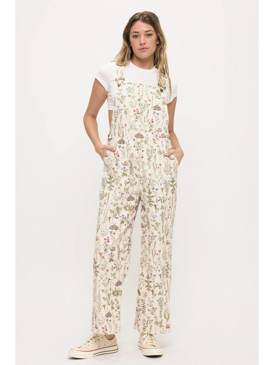 Front view of a woman wearing cream-colored wide-leg overalls with a colorful wildflower and moth print, layered over a white t-shirt.