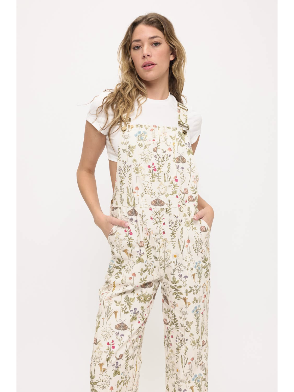 Front view of a woman wearing cream-colored wide-leg overalls with a colorful wildflower and moth print, layered over a white t-shirt.