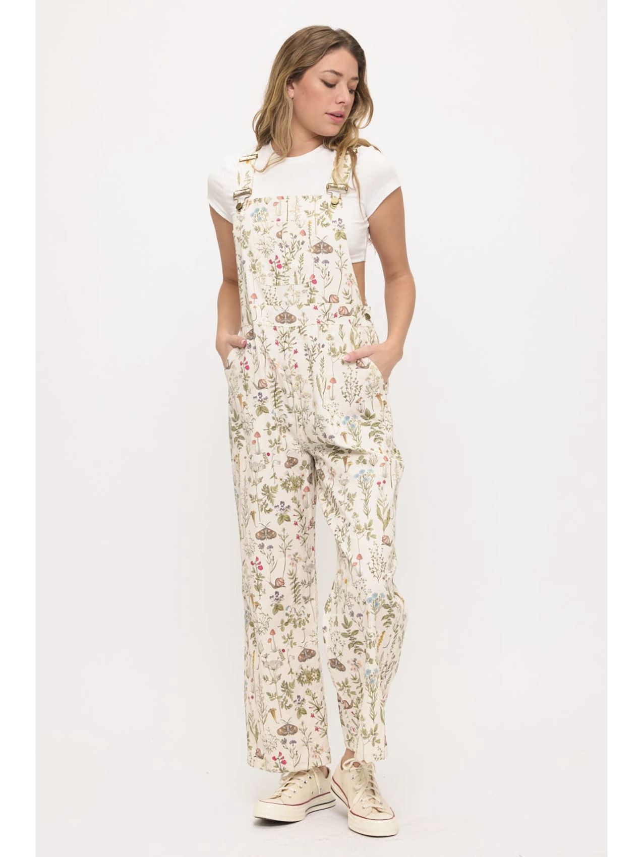 Front view of a woman wearing cream-colored wide-leg overalls with a colorful wildflower and moth print, layered over a white t-shirt.