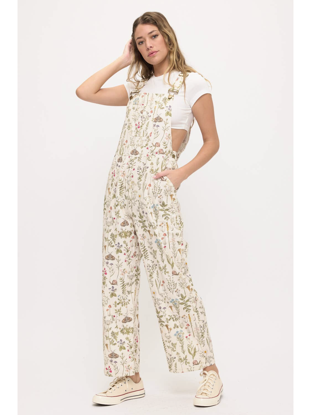 Front view of a woman wearing cream-colored wide-leg overalls with a colorful wildflower and moth print, layered over a white t-shirt.