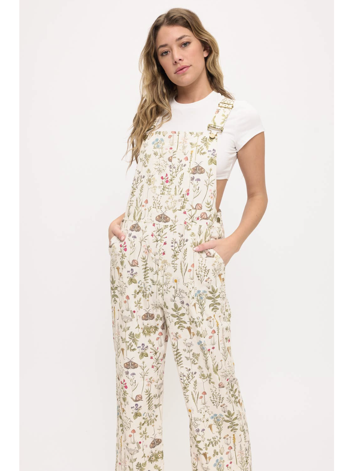 Front view of a woman wearing cream-colored wide-leg overalls with a colorful wildflower and moth print, layered over a white t-shirt.