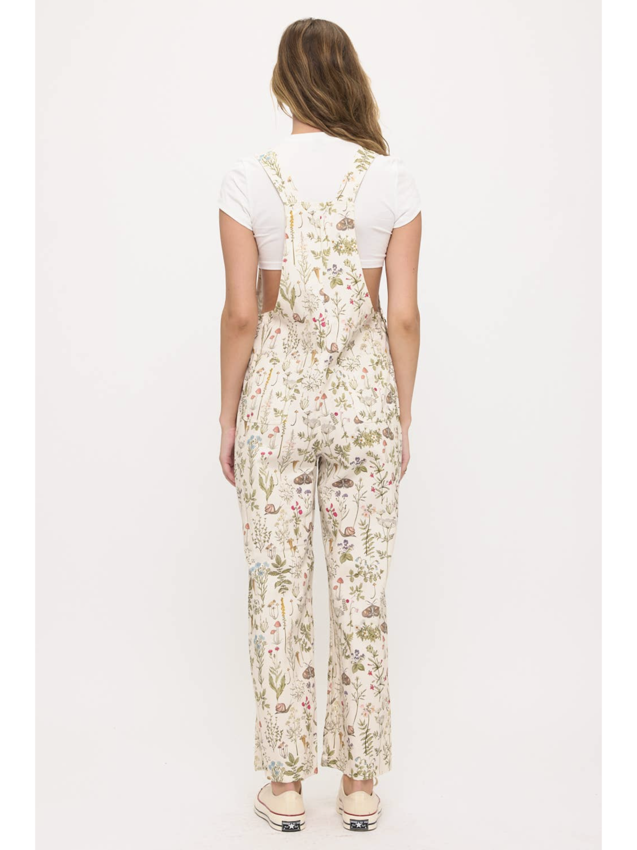 Back view of a woman wearing cream-colored wide-leg overalls with a colorful wildflower and moth print, layered over a white t-shirt.