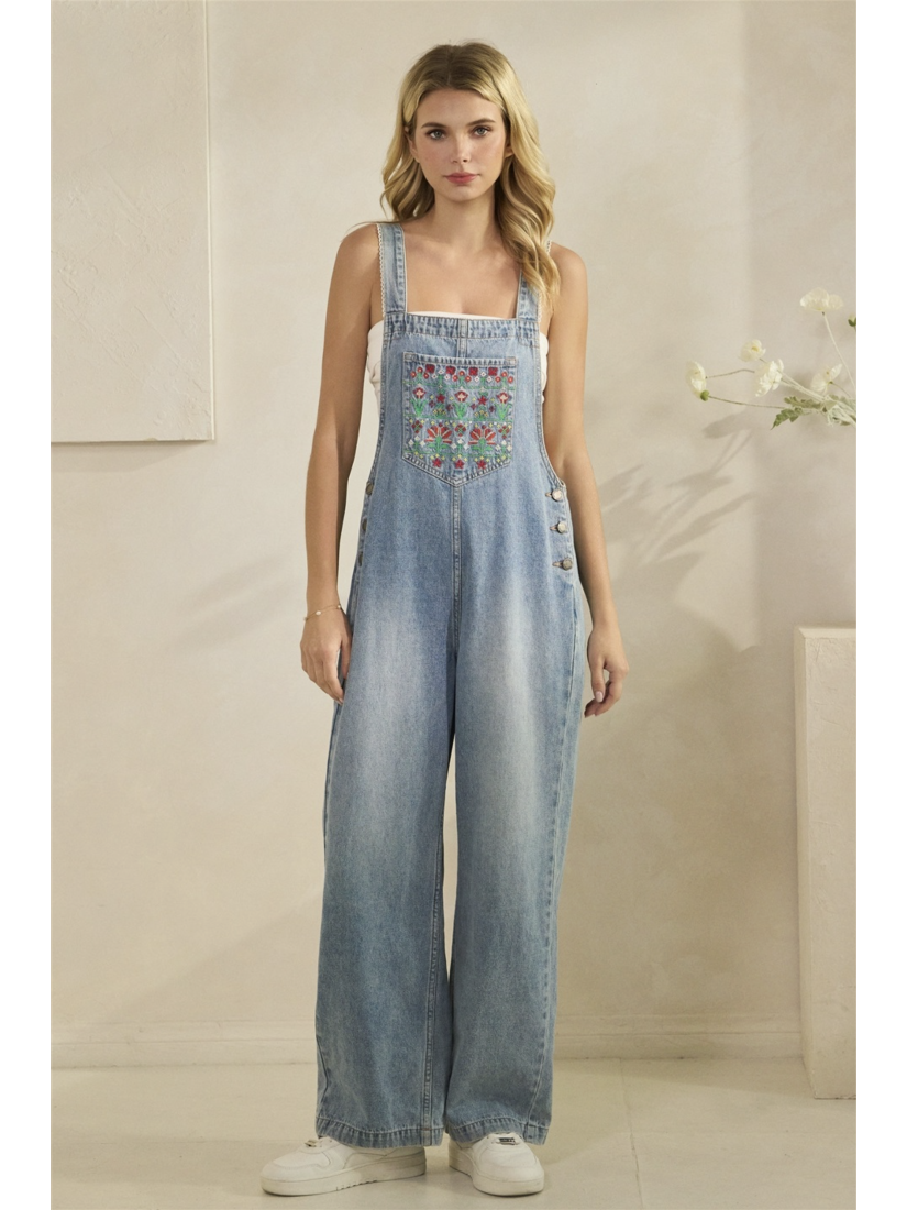 A woman wearing mid-wash blue denim overalls with colorful floral embroidery on the front bib pocket and leg, standing against a neutral background.