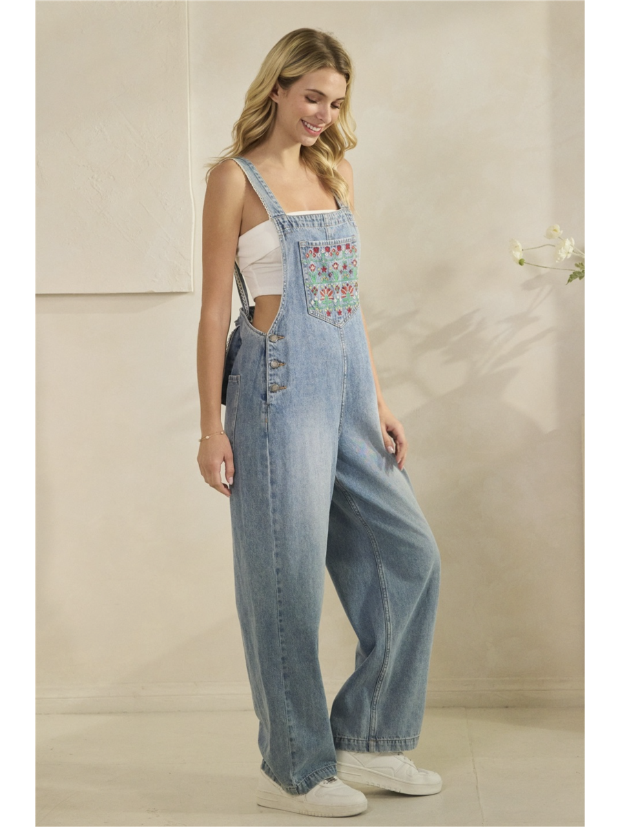 A woman wearing mid-wash blue denim overalls with colorful floral embroidery on the front bib pocket and leg, standing against a neutral background.