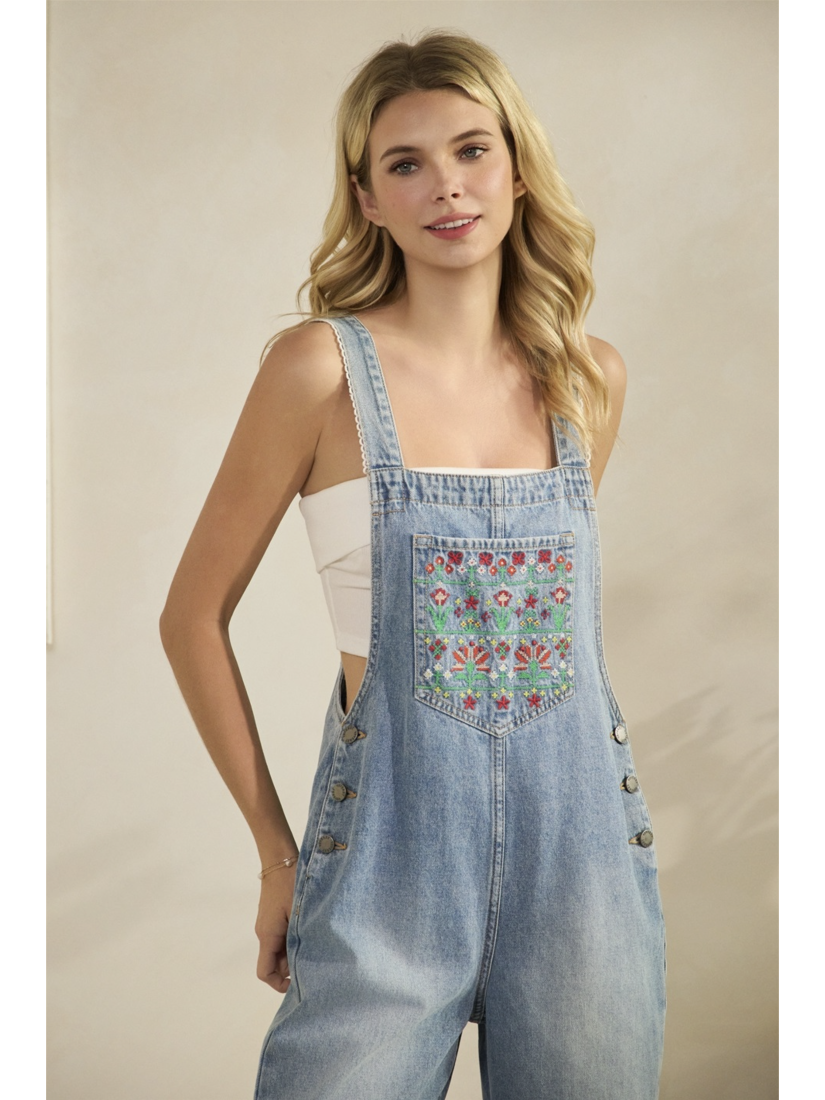 A woman wearing mid-wash blue denim overalls with colorful floral embroidery on the front bib pocket and leg, standing against a neutral background.