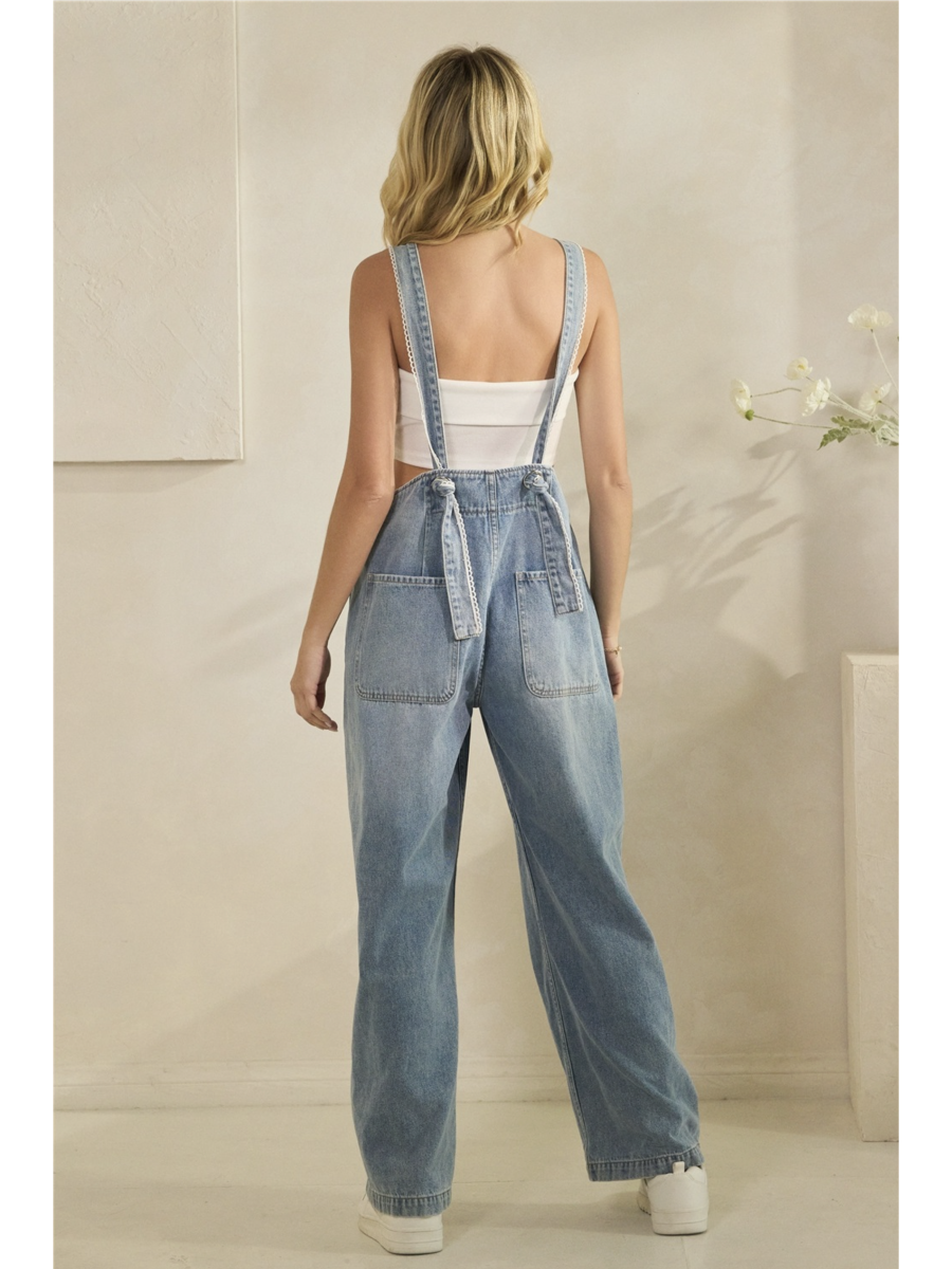 A woman wearing mid-wash blue denim overalls with colorful floral embroidery on the front bib pocket and leg, standing against a neutral background.