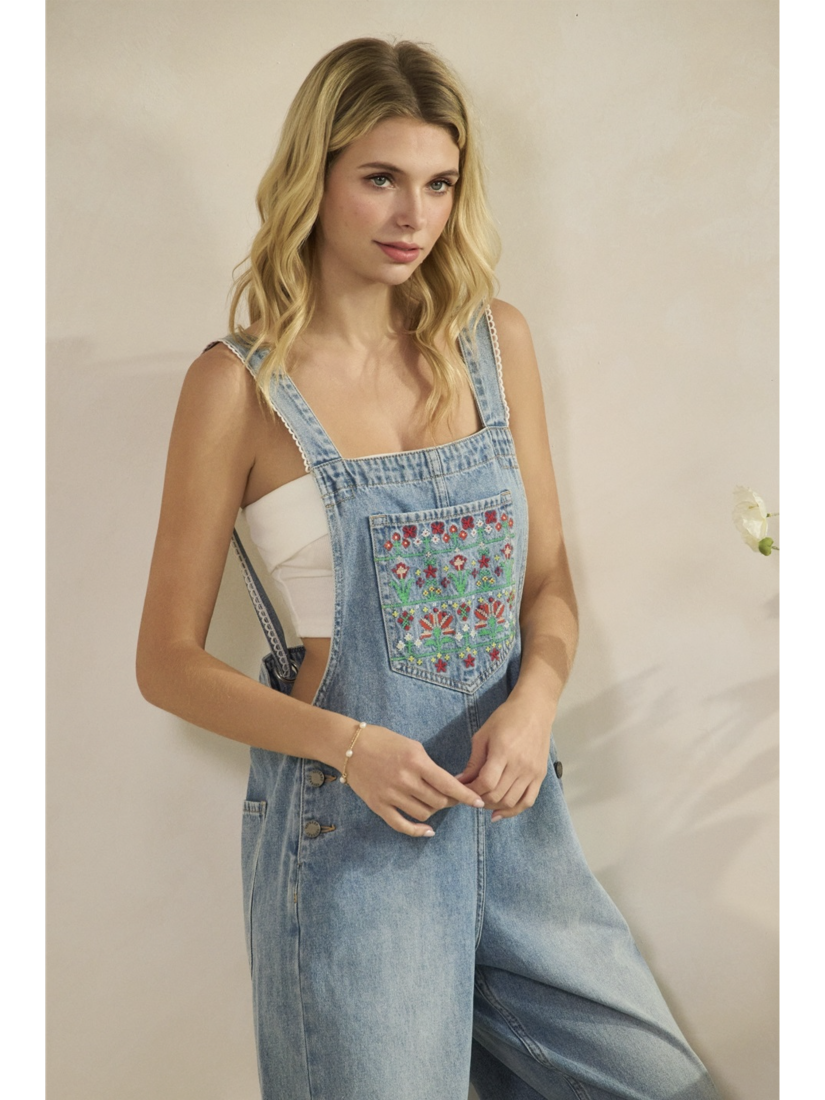 A woman wearing mid-wash blue denim overalls with colorful floral embroidery on the front bib pocket and leg, standing against a neutral background.
