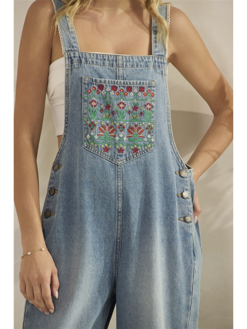 A woman wearing mid-wash blue denim overalls with colorful floral embroidery on the front bib pocket and leg, standing against a neutral background.