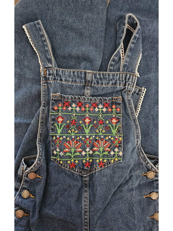 A woman wearing mid-wash blue denim overalls with colorful floral embroidery on the front bib pocket and leg, standing against a neutral background.