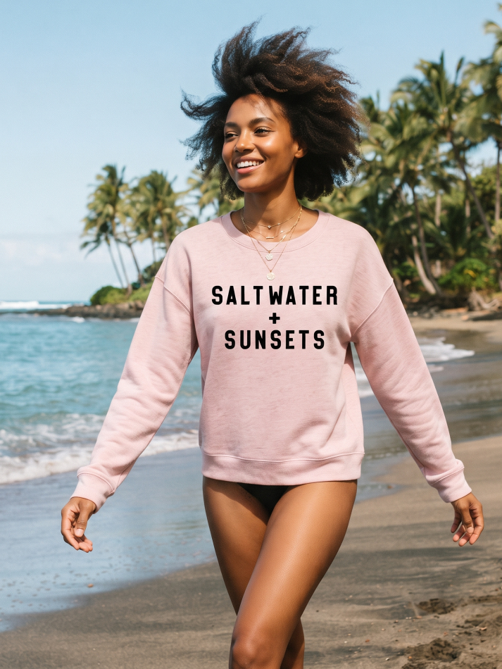 A woman walking on a sandy beach wearing a light pink crewneck sweatshirt with the text 'SALT WATER + SUNSETS' printed in black across the chest.