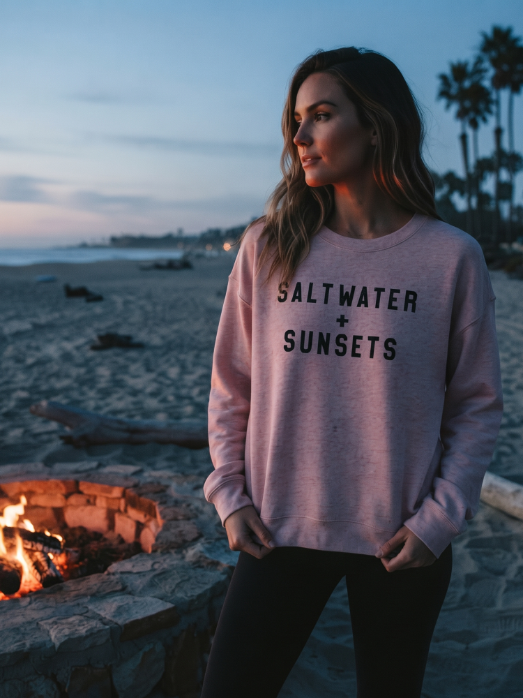 A woman stands on a beach at sunset wearing a light pink heather sweatshirt with the text 'SALT WATER + SUNSETS' printed in black across the chest, paired with black pants. A stone fire pit with flames is visible in the foreground, and palm trees and the ocean are in the background.