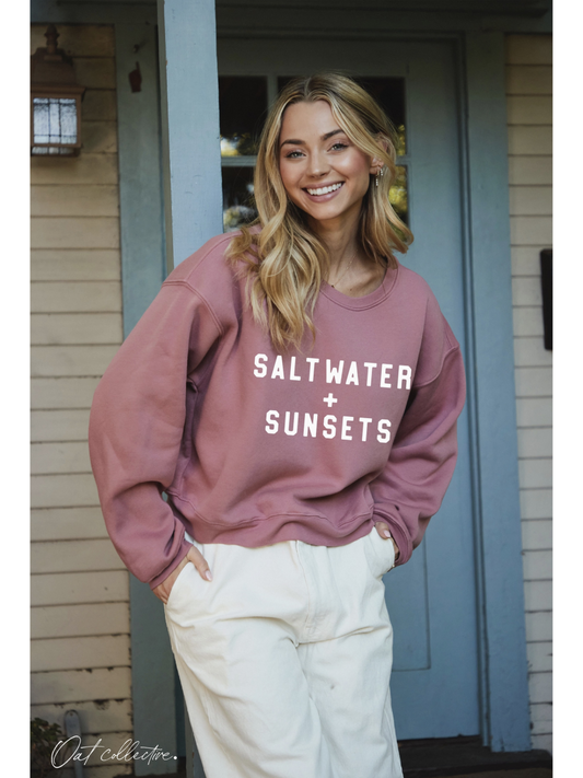Woman wearing a pink sweatshirt with 'Saltwater + Sunsets' text, standing in front of a house.