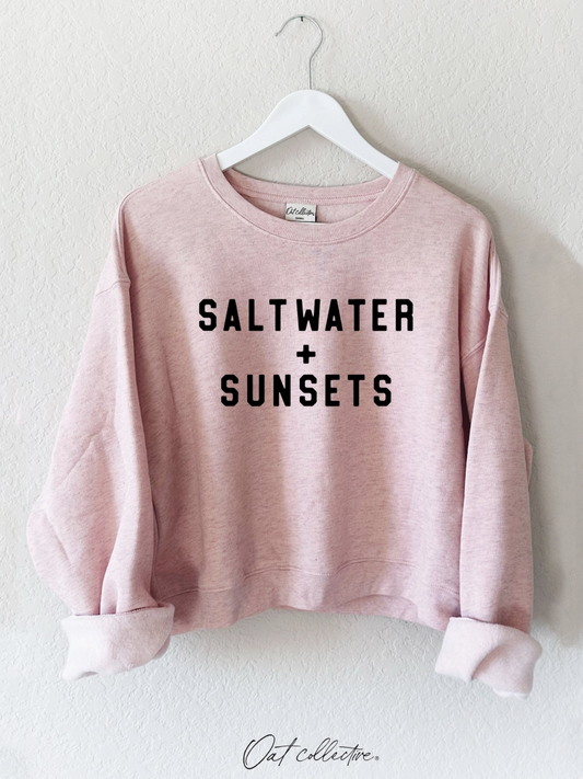 Pink sweatshirt with 'Saltwater + Sunsets' text on a hanger against a white background