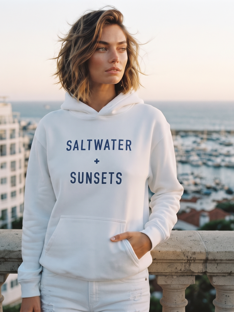 A woman wearing a white pullover hoodie with 'SALTWATER + SUNSETS' printed in navy blue on the front, standing on a balcony with a coastal marina and ocean in the background.