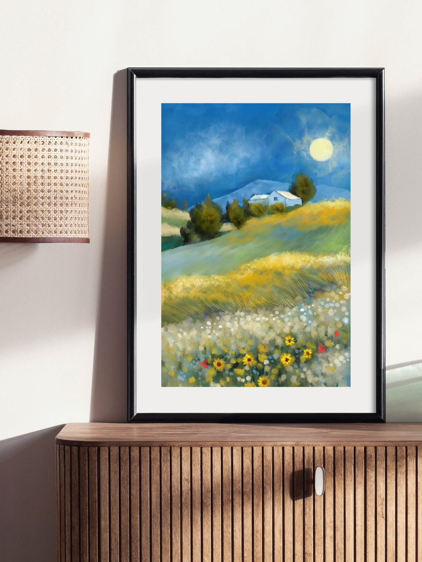 A framed landscape art print leaning against a wall on a wooden cabinet. The artwork depicts a vibrant countryside scene with rolling hills, a white house, trees, a bright moon, and a field of flowers under a deep blue sky.
Artwork by @thomas.little.design