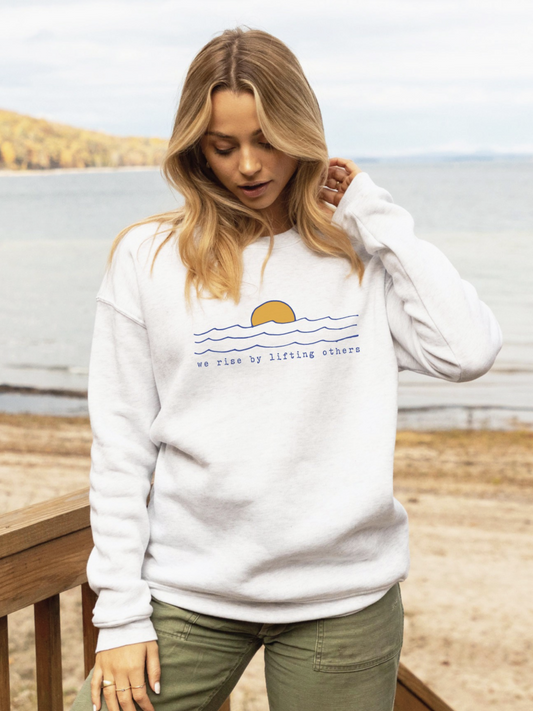 Cozy sweatshirt with a whimsical "We Rise By Lifting Others" graphic, featuring a soft-brushed texture and a relaxed, soulful aesthetic.