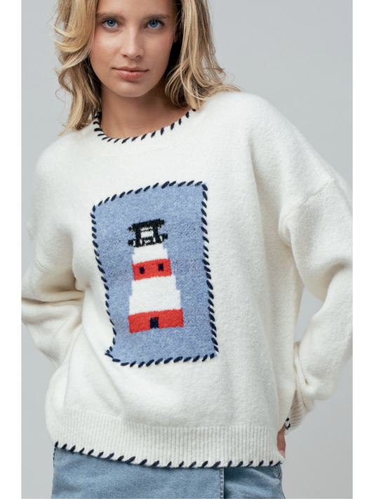 A woman wearing a cream knit sweater featuring a blue and white lighthouse design and blue whipstitch trim on the collar, cuffs, and bottom hem.