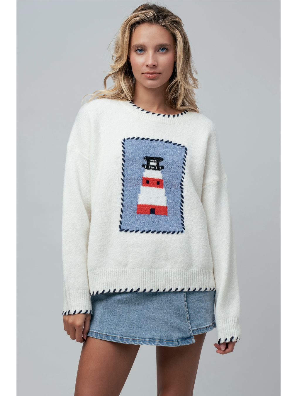 A woman wearing a cream knit sweater featuring a blue and white lighthouse design and blue whipstitch trim on the collar, cuffs, and bottom hem.