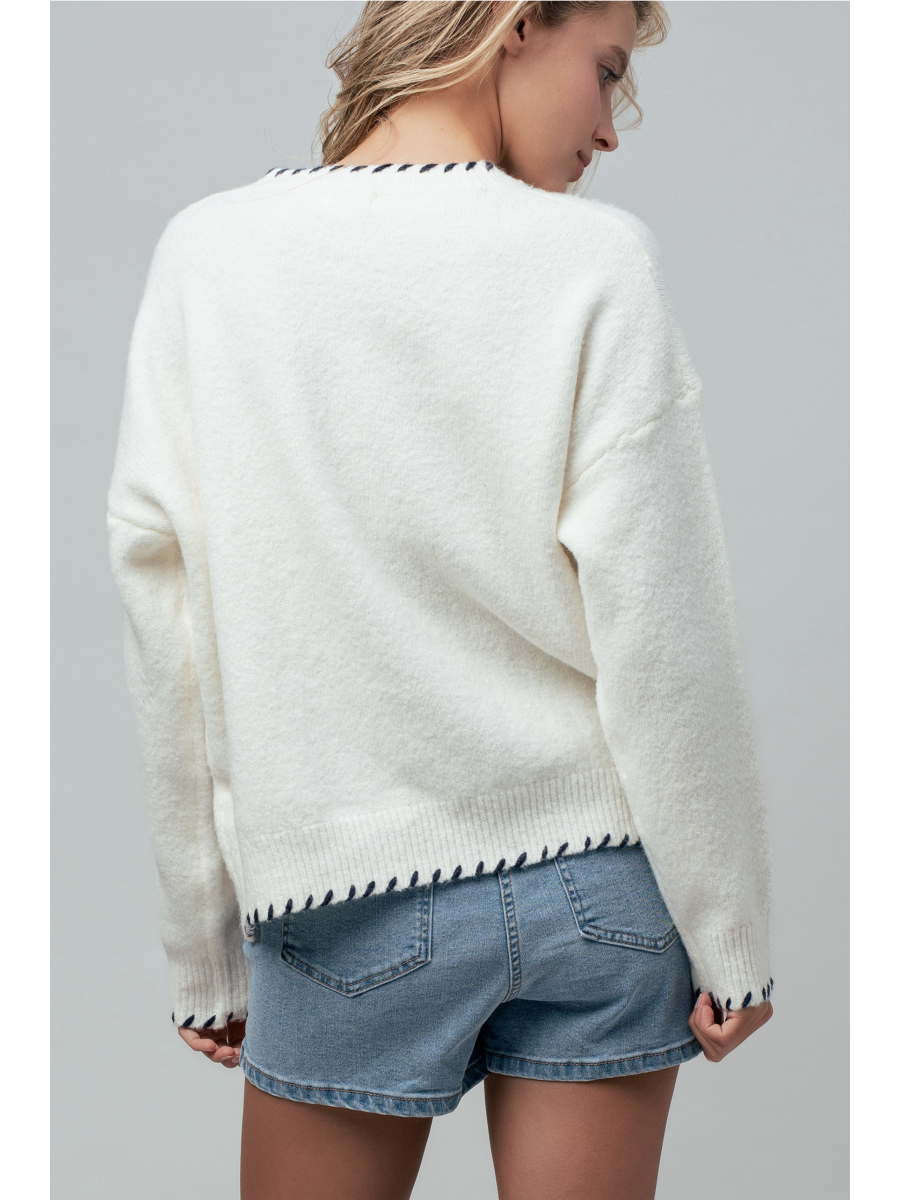 A woman wearing a cream knit sweater featuring a blue and white lighthouse design and blue whipstitch trim on the collar, cuffs, and bottom hem.