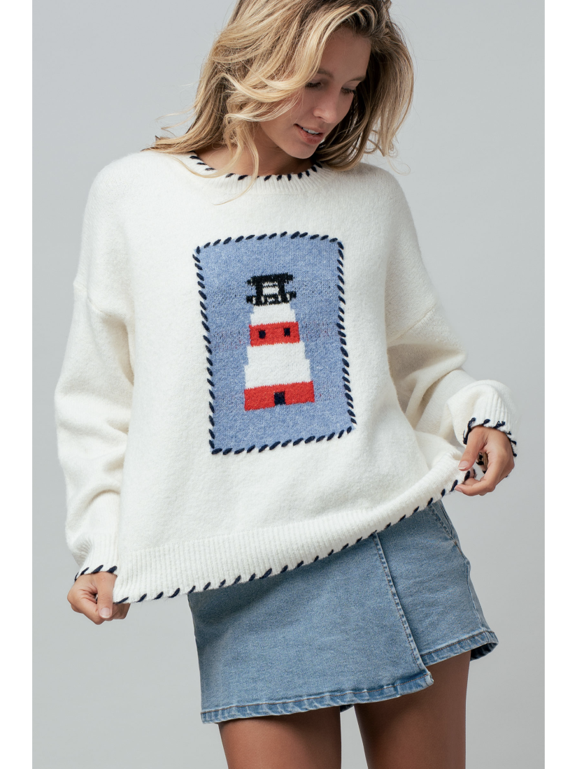 A woman wearing a cream knit sweater featuring a blue and white lighthouse design and blue whipstitch trim on the collar, cuffs, and bottom hem.